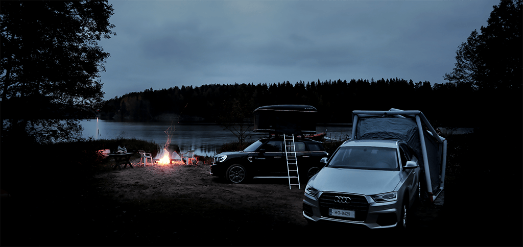 Audi Q3 ja Mini Countryman järven rannalla iltahämärässä. Nuotioon lisätään puuta ja kipinät lentävät taivaalle.